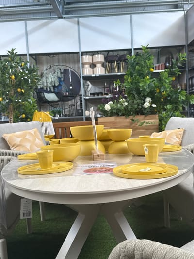 The image depicts an indoor setting, likely a store, displaying outdoor furniture and decor. In the foreground, a round, light-colored table is set with a yellow dinnerware set, including plates and cups. The table is covered with a clear plastic sheet. Behind the table, several yellow bowls are stacked. To the left, a gray armchair with a yellow patterned cushion is visible. Further back, a faux lemon tree with yellow fruit adds a touch of greenery. In the background, shelves are stocked with various items, including decorative vases, planters, and other home goods. The overall environment suggests a retail space, possibly a garden center or home furnishings store, located in Tuk, Netherlands. There are no people visible in the image.