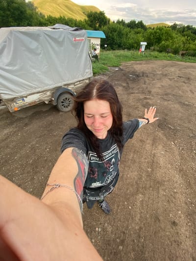 In Верхнеозёрное, Russia, a young woman with long brown hair, wearing a dark t-shirt with a graphic print and patterned sneakers, is taking a selfie outdoors on a dirt ground. She has a tattoo on her left arm and is wearing a silver bracelet. To her left is a grey-covered trailer. The background shows green trees and rolling hills under a cloudy sky. A small building with a blue roof is also visible.