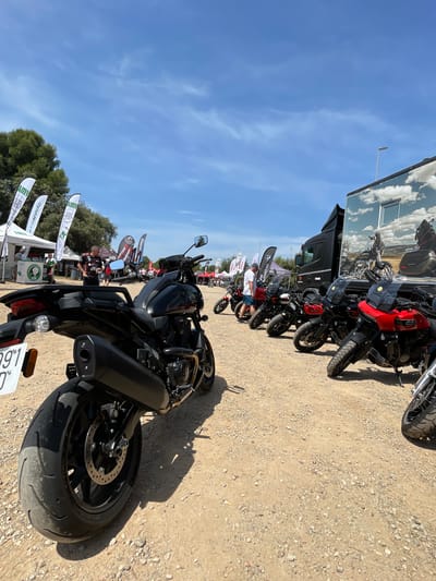 The image depicts a row of motorcycles parked on a gravel lot under a blue sky in l'Ametlla del Vallès, Spain. A large black truck with a cloud-printed advertisement is parked nearby. Several white flags with logos stand near the edge of the lot. People are milling about in the background. The foreground features a black motorcycle with a visible license plate.