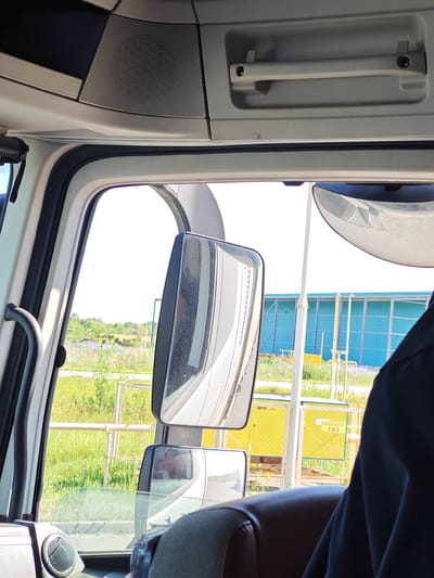 The image is taken from inside the cab of a truck in Chapaevsk, Russia. Through the side window, a landscape of green grass and trees is visible, with a bright blue building in the background. There are two mirrors visible from the interior of the truck, one large exterior mirror reflecting the sky and a blue building, and a smaller interior mirror reflecting a person's face. A black fabric is draped over the passenger seat in the foreground. The truck's interior features a speaker grille on the ceiling and a gray handle above the windshield. Yellow signs with Cyrillic text are visible outside the truck, near a metal fence. The overall scene suggests a daytime setting on the outskirts of a town or industrial area.