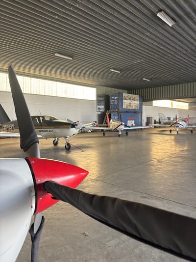 The scene depicts the interior of an aircraft hangar at Toussus-le-Noble, France. In the immediate foreground, the red spinner and a dark grey-sleeved propeller blade of an aircraft are prominently featured on the lower left. The hangar floor is grey concrete.

Several light aircraft are parked inside the hangar. To the left, a white aircraft with black stripes, bearing the registration F-GRBW and the text "AERO-CLUB AIR FRANCE II," is parked. Further back and to the right, at least three additional white light aircraft, each with red tail accents, are aligned. One of these displays "APL" on its fuselage.

In the background, two blue shipping containers are stacked against a white wall; the lower container clearly displays "APL." The hangar ceiling is composed of corrugated metal, from which linear fluorescent light fixtures are suspended. Large horizontal windows situated high on the walls allow natural light to illuminate the space. No individuals are present, and no active interactions or movements are depicted.