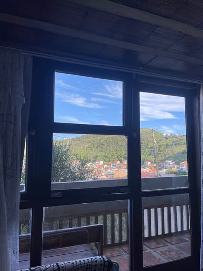 A view through a multi-pane window reveals a bright sky and a distant hillside dotted with buildings and foliage. The window's dark frame contrasts with the light outside. To the right, a portion of a balcony with wooden railings is visible. The interior wall on the left has a textured, patterned surface, and a white, possibly lacy, fabric drapes along the bottom of the window.