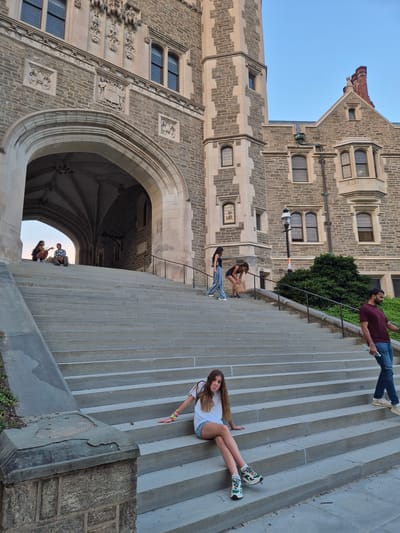 The image depicts a wide, stone staircase leading up to an arched entryway of a large, gothic-style stone building, likely on the Princeton University campus in Princeton, New Jersey. The building features intricate stone carvings above the archway and numerous windows. Several people are present on the stairs. In the foreground, a young woman with long brown hair is sitting cross-legged on a step, wearing a white t-shirt and denim shorts. To her right, a man in a burgundy t-shirt and jeans walks down the stairs, holding a water bottle. Further up the stairs, two women are walking down, one in a black tank top and patterned pants, the other in a black tank top and denim shorts, carrying a paper bag. Near the top of the stairs, under the archway, two people are sitting and talking. The sky is clear and blue, suggesting it is late afternoon or early evening. Lush green bushes are visible on the right side of the image.