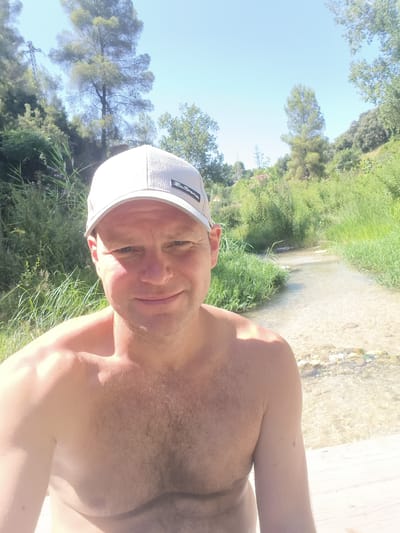 A shirtless man wearing a light-colored baseball cap is posing for a selfie outdoors. He has short hair and is smiling. Behind him, a stream flows through a natural environment with lush green vegetation and trees under a clear blue sky. The setting appears to be a park or natural reserve. This scene is located in the city of Ontinyent, Spain.