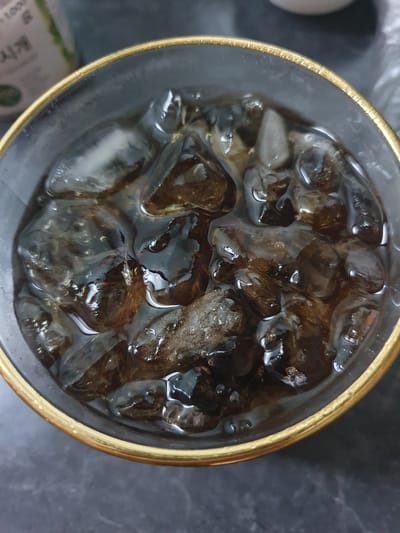 A close-up, overhead shot shows a glass filled with iced coffee. The glass has a gold rim and appears to be sitting on a dark, textured surface. Ice cubes, some partially submerged in the dark liquid, are visible throughout the glass. Small bubbles are present on the surface of the liquid. In the blurred background, a portion of a label from a bottle or container is visible with Korean text that appears to read "시계" (sih-gye), meaning "watch" or "time." The location of Gimhae-si, South Korea is not discernible from the image.