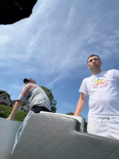 The image depicts two men on a boat, with a section of a quilted gray boat seat in the foreground. One man, on the right, is wearing a white t-shirt with the "Converse Chuck Taylor All Star" logo and white shorts. He is looking upwards and to his left. The second man, on the left, is wearing a grey t-shirt with a flame graphic and blue jeans. He is also looking away from the camera, towards the left. Behind them, the sky is blue with scattered clouds, and a green grassy embankment with houses and trees is visible. The scene appears to be outdoors, likely on a body of water near Gellicum, Netherlands.
