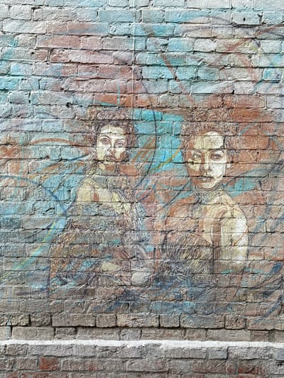 The image shows a mural painted on a brick wall in Zhabkino, Russia. The mural depicts two women in what appears to be a stylized, artistic representation. The women are rendered with muted colors and a textured, almost sketch-like quality, integrated into the rough surface of the bricks. The background of the mural features abstract shapes and lines in blues, browns, and hints of orange. The brick wall itself is weathered and aged, showing variations in color and texture, with some bricks darker and others lighter. A horizontal ledge or shelf made of lighter-colored material runs across the bottom of the image, with more bricks visible below it. The overall impression is one of urban art on an old structure.