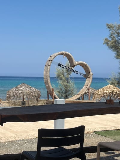 In Protaras, Cyprus, a large woven heart sculpture is positioned on a sandy beach, framing the turquoise waters of the Mediterranean Sea. The sculpture, made of rope, has the word "MONTEGO" emblazoned across it, with smaller text below reading "PROTARAS CYPRUS." Two thatched umbrellas offer shade on the beach, and a sign with the number "40" is visible near one of them. In the foreground, a dark chair sits at a wooden table, suggesting an outdoor seating area. The scene is bathed in bright sunlight under a clear blue sky.