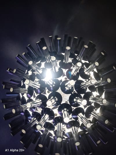 This is a close-up, low-angle, dimly lit photograph of a modern spherical light fixture. The fixture appears to be composed of numerous metallic, cylindrical tubes radiating outwards from a central point, resembling a sea urchin or a dandelion seed head. The tubes are dark, possibly black or deep grey, with a reflective surface that catches the light. Each tube has a circular opening at its end, through which a bright, white light emanates, suggesting individual light sources within or behind them. Some of the tubes show faint purple and blue reflections, indicating the color of the ambient light or the interior of the fixture. The background is a dark, out-of-focus void, emphasizing the intricate detail of the light fixture. There is no visible context of the city of Улукаўскі сельскі Савет, Belarus within the image itself. A watermark in the bottom left corner reads "A1 Alpha 20+".