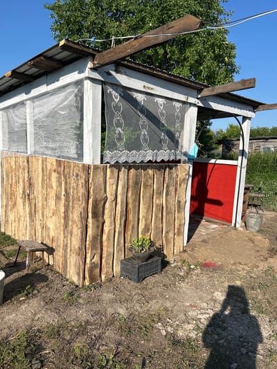 The image depicts an outdoor scene in Покров ауылдық округі, Kazakhstan, featuring a rustic, partially constructed open-air structure. The structure has a corrugated metal roof supported by wooden beams, some of which appear weathered and unfinished. The walls are a combination of white-painted vertical planks and a lower section constructed from rough-hewn, unprocessed logs, giving it a raw, natural aesthetic. Delicate white lace curtains hang across parts of the structure, offering a stark contrast to the rugged exterior.
To the right of the structure, a vibrant red door is visible, its color standing out against the muted tones of the surrounding environment. The ground is uneven, composed of dirt, gravel, and patches of dry grass, suggesting a rural or semi-developed setting. Various objects are scattered around: a small wooden stool sits to the left, a black plastic crate containing a potted plant with yellow flowers is placed near the log wall, and a metal bucket is situated near the red door.
In the foreground, a distinct shadow of a person holding a camera or phone is cast on the ground, indicating the photographer's presence and perspective. In the background, lush green trees and foliage are visible, along with glimpses of other rustic structures, reinforcing the rural character of the location. The sky is clear and blue, suggesting a sunny day. The overall impression is one of a casual, perhaps ongoing, outdoor building project in a natural, rural setting.