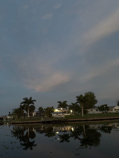 The image depicts a serene canal scene at dusk in North Port, Florida, USA. The water is calm, reflecting the silhouettes of palm trees and houses along the embankment. Several single-story houses with screened porches are visible, some illuminated by interior lights. A wooden dock extends into the water on the left, with a small boat moored to it. Another dock is present in the mid-ground, also with a boat. The sky is a muted twilight blue with wispy clouds, hinting at the transition from day to night. The overall atmosphere is peaceful and residential, characteristic of a waterfront community.