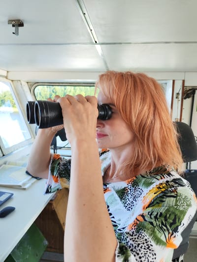 A woman with shoulder-length, reddish-blonde hair is looking through a pair of black binoculars. She is inside a room that appears to be the control cabin of a vessel, with a window to her left offering a view of trees and water. The woman is wearing a brightly colored, patterned shirt with abstract designs of leaves and animal prints in green, orange, white, and black. She has a silver necklace on.
In the background, a desk is visible with a computer mouse and what appears to be a navigation chart or map. A black headrest of a chair is also present to the right. The room has a white ceiling with metal fixtures. The lighting suggests it is daytime, and the weather appears clear. The overall scene evokes a sense of vigilance and navigation. No visible text is present. The location context is Даргун, Russia.