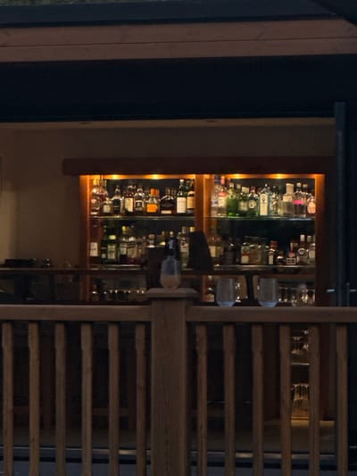 The image displays an evening view of a bar area, observed from an outdoor or semi-outdoor vantage point in Bubwith, United Kingdom. In the immediate foreground, a wooden railing with vertical balusters and a flat top rail spans the width of the frame. A light-colored, stemless glass, appearing to contain a beverage, rests on a prominent wooden post of this railing on the left. Two additional clear, empty stemless glasses are positioned on the railing to the right.
Behind the railing, a well-appointed bar counter is visible. Above the counter, a large, illuminated display cabinet is recessed into a wall, showcasing a comprehensive collection of liquor bottles. These bottles, varying in size, shape, and color, are neatly arranged on multiple glass shelves within a wooden frame. Warm, amber-toned lighting within the cabinet highlights the bottles, which include various spirits such as gins, vodkas, whiskies, and liqueurs. A dark, indeterminate object rests on the bar counter to the far left. The area above and behind the liquor display is dark, suggesting an overhead structure or night environment. No individuals are present in the scene, and no specific actions or interactions are depicted.
