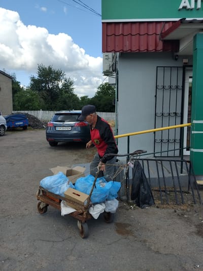 In Chudniv, Ukraine, a man wearing a dark baseball cap, a red and black jacket, and grey pants is pushing a rusted, four-wheeled cart loaded with blue plastic bags and cardboard boxes. The cart is positioned on a cracked asphalt surface in front of a grey building with a red tiled roof. A yellow handrail runs along the building's exterior, next to a black metal gate. Behind the cart, a dark grey car with the license plate "AM 5649 EM" is parked. In the background, a white picket fence separates the parking area from a wooded area and other buildings. The sky above is blue with scattered white clouds.