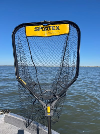 A fishing net, branded "SPORTEX GERMANY", is positioned in the foreground, obscuring the view of a body of water. The net's frame is black with a yellow label at the top. The net itself is black mesh. The water appears choppy under a clear blue sky. The lower edge of a boat, made of aluminum, is visible at the bottom of the frame. The location is specified as the city of Novopichugovo, Russia, however, this information is not visually verifiable from the image alone. There are no people or animals present in the frame.