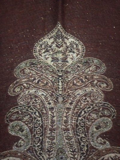 A brown textile with a large, ornate, paisley-like design woven into the fabric. The design consists of repeating, curvilinear motifs with intricate detailing and embellishments in shades of silver, gold, and black. The paisley elements are arranged vertically, forming a symmetrical, almost tree-like pattern. The background brown fabric appears to have a subtle, shimmering quality, suggesting the presence of metallic threads or fibers.