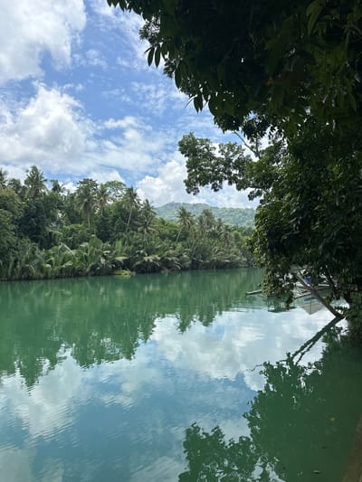 The image depicts a serene river scene in Valladolid, Philippines. The river, a vibrant turquoise green, dominates the foreground, reflecting the sky and the lush vegetation along its banks. Dense tropical foliage, including palm trees and other verdant plants, lines the river, creating a sense of natural abundance. In the background, rolling hills covered in greenery suggest a mountainous or hilly terrain. The sky above is a bright blue, dotted with fluffy white clouds, contributing to the overall tranquil atmosphere. A small boat, possibly a banca, is partially visible near the right bank, hinting at local transportation or leisure activities. The water appears calm, with subtle ripples that distort the reflections of the sky and trees. The overall impression is one of natural beauty and peacefulness in a tropical setting.