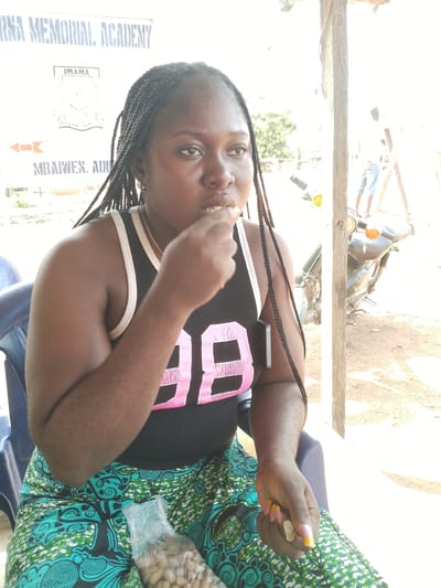 In Ichol, Nigeria, a young woman with braided hair is seated outdoors, her gaze directed downwards as she brings food to her mouth. She wears a black tank top with pink "98" graphics and a vibrant green and teal patterned skirt. Her hands are adorned with long, yellow and pink manicured nails and rings. A clear plastic bag filled with nuts rests beside her on her lap, near her skirt. The background reveals a wooden post, a motorcycle, and a sign that reads "IMAMA" and "MBAWLEN. ADI". A distant figure stands near a wooden structure. The lighting suggests it is daytime.