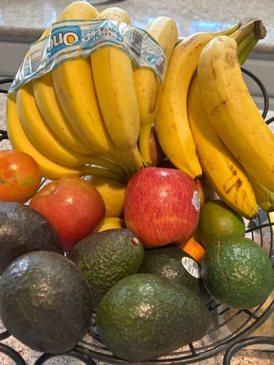 The image presents a close-up, high-angle view of a black metal wire fruit basket filled with various fresh produce. A large cluster of ripe yellow bananas occupies the top and center, with some bananas still bound by clear plastic packaging displaying "One" branding. Several individual bananas show minor brown spots. Beneath and around the bananas are multiple red apples, one with a white PLU sticker bearing the number 4133. Scattered throughout the lower portion of the basket are numerous dark green to blackish-green avocados with textured skin. A bright green lime is visible towards the right, and a reddish-orange tomato is on the far left. A partial view of an orange-colored citrus fruit is also nestled among the other items. The fruit basket rests on a light-colored, speckled countertop, likely granite, with a blurred background suggesting an indoor domestic kitchen environment. No individuals are present, and the scene depicts a static arrangement of items.