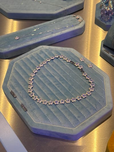 The image shows a display of jewelry, likely in a store. Two necklace displays, covered in light blue, are visible. The one on the right has a necklace on it with floral, sparkling stones set closely together in a ring. To the left is a second, similar display with a pendant necklace. The background is a mottled gold color. The presence of jewelry in a showcase suggests this image was taken inside a jewelry store. I do not have enough information to determine the location in Spreitenbach, Switzerland.