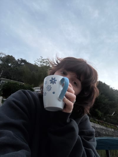 The image captures an individual holding a white ceramic mug with a light blue handle and blue floral designs. The person has dark brown, mid-length hair with bangs and wears a dark-colored long-sleeved garment. Their eyes are directed towards the camera, while the mug partially obscures the lower portion of their face. A ring is visible on the right hand. The background features an overcast sky and a densely vegetated hillside. On the left, a light-colored architectural structure with visible railing sections and retaining walls is present. The scene is outdoors in the city of San Pablo La Laguna, Guatemala.
