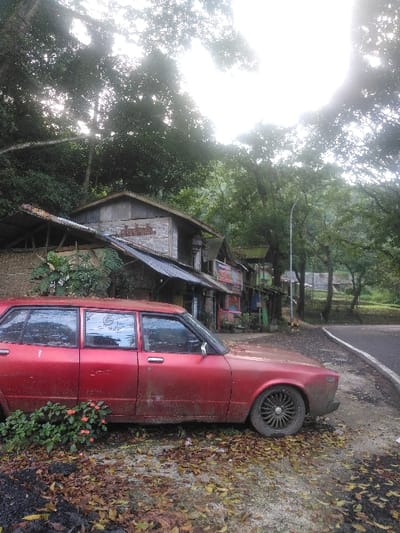 The image captures a static outdoor scene in Cugenang, Indonesia. A dark red, four-door sedan, heavily coated in dirt and grime, is parked on a dirt and leaf-strewn shoulder in the foreground, facing right. The vehicle exhibits faded paint and features multi-spoke black wheels. Small green foliage with red flowers grows directly in front of its left front wheel. Behind the vehicle, a series of aged, single-story structures with corrugated metal roofs and varied construction materials, including wood planks and some brickwork, are visible. Advertisements or posters are affixed to some exterior walls of these structures. Dense green trees and foliage form a significant background on the left and upper frame, with bright sunlight filtering through the canopy, creating distinct lens flare in the upper central and right portions. An asphalt road with a solid white edge line curves to the right of the parked vehicle. A street light pole is positioned between the structures and the road. The ground around the car is covered with accumulated dry leaves and patches of dark earth. No individuals are present in the scene.