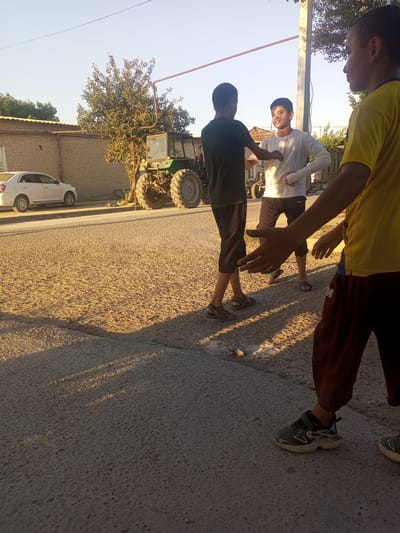 This media file captures a scene in Tahmach, Uzbekistan, during the late afternoon or early evening, as indicated by the long shadows cast across the dusty ground. Three young boys are the primary subjects, engaged in an interaction that appears to be playful or perhaps a friendly dispute. One boy, wearing a dark t-shirt and shorts, faces two others. The boy in the center, dressed in a white t-shirt and dark shorts, has a cast on his right arm and is looking towards the boy in the dark t-shirt, with his left arm extended. The third boy, partially visible in the foreground on the right, wears a yellow t-shirt and dark pants, with his arm outstretched towards the center.
The setting is an outdoor area that appears to be a village or rural roadside. Behind the boys, a weathered brick building with a yellow-tinted roof is visible. Further in the background, a large green tractor is parked, suggesting a connection to agricultural activity. A white car is parked on the left side of the road. The ground is a mixture of asphalt and loose, sandy dirt, characteristic of unpaved roads. The sky is clear and a light blue, with the warm glow of the setting sun illuminating the scene. The overall mood seems casual and perhaps a bit boisterous, given the body language of the boys. There is no visible text in the image.