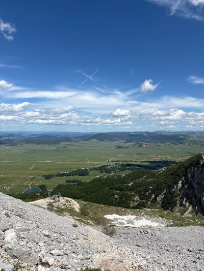 The image is a wide-angle, high-angle shot taken from a rocky, mountainous terrain overlooking a vast, green valley. The sky is a vibrant blue with scattered white clouds, some of which form a distinct 'X' shape. In the foreground, the ground is covered with a dense layer of grey and white rocks, characteristic of a scree slope.

Further down the slope, a small ski lift station is visible, consisting of a building with a green roof and several ski lift towers with red chairs. Adjacent to the ski lift is a small, dark blue lake. Beyond the ski lift and lake, the landscape transitions into a sprawling green valley dotted with scattered houses and fields, indicating a rural settlement. The valley floor is relatively flat and extends towards rolling hills and mountains in the distance, which are layered in shades of blue and green, suggesting depth and distance. The overall scene depicts a natural landscape with elements of human infrastructure, likely a tourist or recreational area in a mountainous region. The location is identified as Pašina Voda, Montenegro.