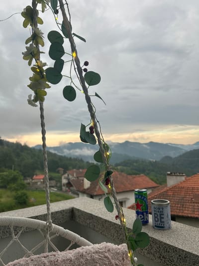 A close-up shot shows a decorative rope with artificial flowers and small lights, hanging in front of a mountainous landscape. On a ledge in the foreground, there are two containers: one is a green can of Heineken beer, and the other is a cylindrical container with text that reads "DON'T WORRY. BE HAPPY." The background reveals a village with red-tiled roofs nestled in a valley, with rolling hills and mountains extending into the distance under a cloudy sky. The location is the city of Ravnishta, Bulgaria.