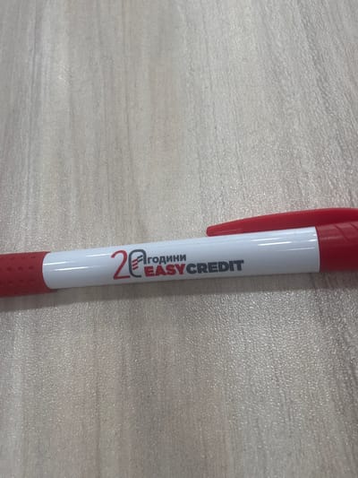 A red and white ballpoint pen rests on a light-colored, textured surface, likely a desk. The pen is positioned horizontally, with the red grip on the left and the white barrel extending to the right. The barrel of the pen features black and red text that reads "20 ГОДИНИ EASY CREDIT." The background is a uniform, lightly textured surface with vertical striations, suggestive of wood grain or a similar material. The lighting is even, with no harsh shadows, indicating an indoor setting. The scene is static and there are no people visible.