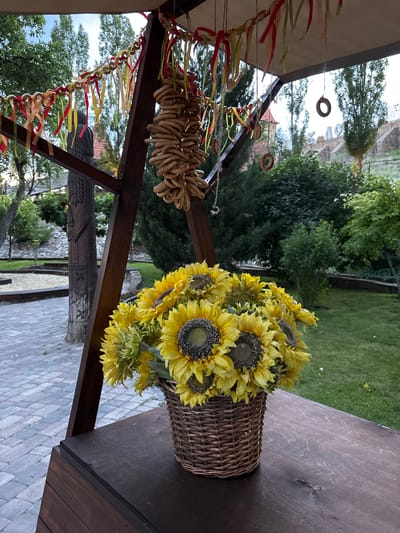 In Staraya Stanitsa, Russia, this image depicts an outdoor setting with a wooden structure that has a canopy. Suspended from the structure are strings of bread rings and colorful ribbons. A basket filled with sunflowers sits on a dark wooden surface. In the background, there is a courtyard with a stone pathway, lush green grass, trees, and a stone wall. A carved wooden post is also visible.