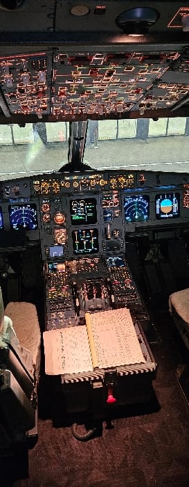 The image displays the interior of an aircraft cockpit, specifically a flight deck, observed from a slightly elevated, central perspective. The location is an airport in Pyrzowice, Poland. The overhead panel is visible at the top, illuminated by numerous orange and white lights on its switches and annunciators. Below this, the main instrument panel features multiple digital displays, including primary flight displays, navigation displays, and engine indication and crew alerting system (EICAS) screens, all actively illuminated with blue, green, and orange graphical information. Analog gauges and various buttons also show illumination. The central pedestal, positioned between two pilot seats (partially visible on the left and right), contains throttle levers and control panels, also illuminated. An open logbook or checklist, containing handwritten text, rests on a surface located on the central pedestal. No individuals are present. Through the cockpit windscreen, an exterior view of what appears to be an airport tarmac or ramp area is visible, with precipitation (rain or snow) evident on the glass, suggesting outdoor conditions. The overall lighting within the cockpit is dim, primarily originating from the instrument panels themselves.