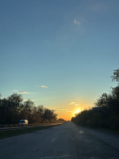 The image captures a road scene at sunset in Korotych, Ukraine. The sun, a bright golden orb, is low on the horizon, positioned centrally to the right, casting a warm glow across the landscape. The sky transitions from a clear, deep blue at the top to lighter blues and vibrant oranges and yellows near the horizon, with a few wispy clouds scattered throughout.
The road, appearing to be asphalt with visible cracks and patches, stretches into the distance, narrowing with perspective. White lane markings are visible. Metal guardrails run along the left side of the road. Dense lines of trees and vegetation flank both sides of the road, many appearing bare or with sparse foliage, suggesting late autumn or early spring. Some of the trees on the left show prominent clusters, possibly mistletoe or nests.
On the left side of the road, behind the guardrail, a white van is visible. The text "O.M.S." is clearly discernible on its side. In the far distance, on the main road, small silhouettes of other vehicles can be seen, heading towards the sunset. On the right side, partially obscured by the roadside foliage, there is a dark, dilapidated concrete or brick structure. The overall impression is one of a serene end-of-day journey through a rural or semi-rural landscape. No people are visible.