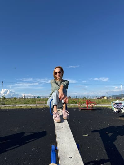 The image features a young woman sitting on a seesaw on a playground. She is wearing a light green jacket, blue jeans, pink sneakers, and sunglasses. The seesaw is made of concrete and metal. In the background, there is a red merry-go-round, a fence, some grass, and buildings. The sky is clear blue with a few white clouds. The sun is shining, suggesting it is daytime. The overall scene is a bright and sunny day at a playground. There is no visible text in the image.