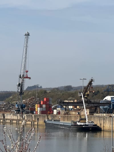 This image depicts an industrial port facility in Mertert, Luxembourg, under an overcast or hazy sky. The scene is dominated by a wide body of water, likely a river, in the lower foreground. A black and white cargo barge named "SUNISHA" is moored alongside a concrete quay wall on the right side of the water.

On the quay, a tall, grey and silver gantry crane equipped with a red container spreader attachment is prominently visible on the left, positioned near a substantial pile of scrap metal. Further to the right along the quay, a second, darker-colored gantry crane with a large grab bucket is present, indicating bulk material handling capabilities. Behind the quay, a large quantity of varied scrap metal forms a significant pile. Adjacent to this, a tall stack of multi-colored shipping containers (red, yellow, grey, white) stands. Industrial structures, including light poles with large luminaires, and additional piles of materials are observed across the quay. In the distant background, hills covered with bare and budding trees are visible. The immediate foreground, out of focus, features a branch with small white blossoms. No individuals are discernible in the scene.