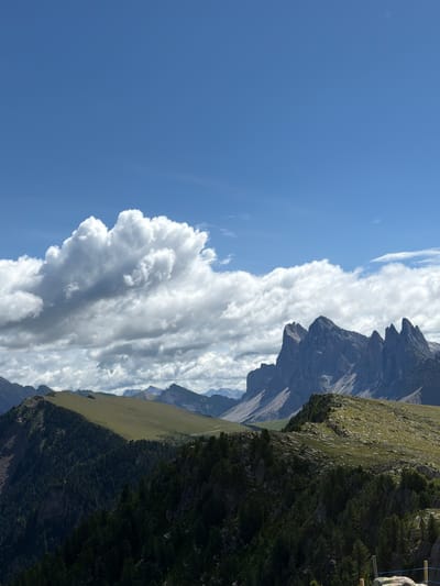 This image depicts a panoramic view of the Dolomites in Ortisei, Italy, on a clear day with scattered clouds. The foreground is dominated by dark green pine forests blanketing steep slopes. In the midground, a grassy ridge curves from left to right, transitioning to a rocky, uneven terrain dotted with green vegetation. In the background, jagged, dark grey mountain peaks pierce the sky, their sharp edges contrasting with the softer contours of the lower slopes. The sky above is a vibrant blue, with large, fluffy white clouds clustered on the left and a few smaller, wispy clouds stretched across the horizon. The scene captures the rugged natural beauty of the mountainous region.