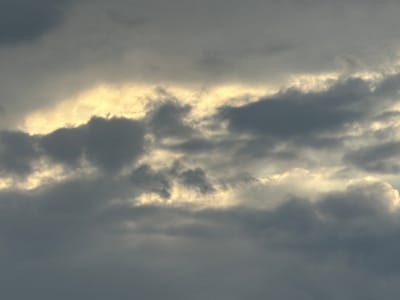 The image captures a dramatic sky filled with dense, dark clouds that are partially illuminated by a bright, golden light breaking through them. The light creates a band across the middle of the frame, highlighting the edges of the clouds and suggesting either sunrise or sunset. The overall mood is somber and atmospheric. The image does not depict any specific urban environment, nor does it contain any recognizable landmarks. There are no people, subjects, objects, or actions depicted in this image. The scene is purely a natural phenomenon of the sky.