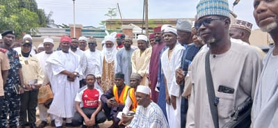 A group of men and one woman are gathered outdoors, with a building and trees in the background. The group appears to be diverse in age, and most of the men are wearing traditional Islamic attire, including various styles of caps and robes. The woman is wearing a uniform.

Several men are wearing patterned caps, and some have beards. One man in the foreground is wearing glasses and a light-colored robe. He has a dark strap across his chest and a phone in his pocket. Another man is wearing a red and white football jersey with the text "Emirates FLY BETTER" visible. Some individuals are holding objects, including what appears to be a prayer mat and a bag.

The scene suggests a social gathering or a formal event. The lighting indicates it is daytime, and the presence of trees suggests an outdoor location. The weather appears to be clear. The location context is Sabon Gari North, Nigeria. There are no discernible emotions clearly expressed by the individuals in the image.