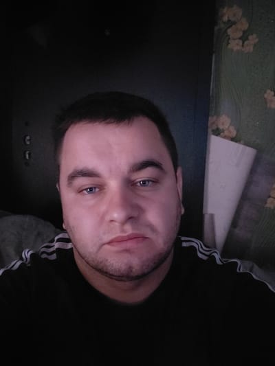 The image is a selfie taken by a man with blue eyes and dark hair. He is wearing a black t-shirt with white stripes on the shoulders. The background is dark, with a wall covered in floral wallpaper on the right side. There is also a large white object that appears to be a roll of paper or fabric in the background. The scene appears to be indoors. The exact location within Ketrysanivka, Ukraine, cannot be determined from the image alone.