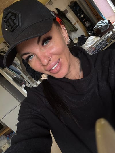 A woman, wearing a black baseball cap and a black hooded sweatshirt, is taking a selfie indoors. She has blue eyes, a nose ring, and visible eyelashes. Her hair is pulled back in a ponytail. The background appears to be a cluttered room with shelves filled with various items, including clothing and household goods. The lighting suggests it is daytime. There is no identifiable information about the location being Dulepovo, Russia.