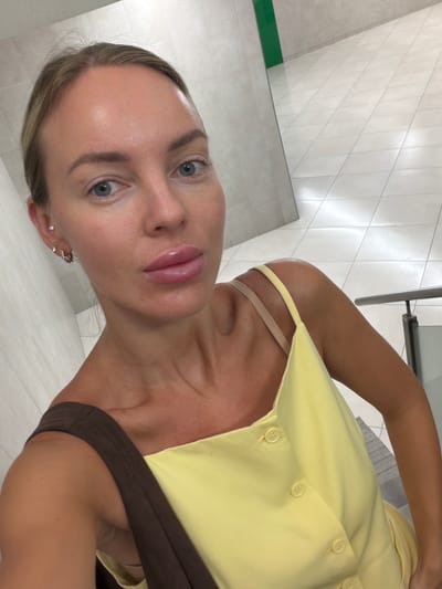 A woman with blonde hair pulled back and blue eyes is taking a selfie in a brightly lit, modern building with tiled floors and white walls. She is wearing a yellow top with thin straps and a brown strap over her shoulder. The background shows a staircase with a metal railing and a reflection of the woman and the environment in a mirror. The location is identified as a city in Spain, specifically Villajoyosa/la Vila Joiosa.