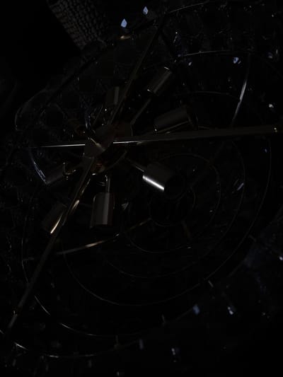 The image displays a dark, intricate metallic structure, possibly a chandelier or abstract sculpture, positioned against a black or deeply shadowed background. A central hub of brass or gold-toned metal anchors multiple radiating arms and cylindrical components. These components exhibit polished, reflective surfaces, catching ambient light to create distinct highlights and glints. Surrounding and partially obscured by the central structure are several concentric or spiraling bands of dark metal. In the upper left quadrant, a subtly textured surface with a hexagonal or honeycomb pattern is visible, suggesting a ceiling or a larger decorative element. The scene is devoid of people, and no specific actions or interactions are depicted. The environment is characterized by deep shadows and metallic reflections. The location in the city of Griboyedov, Armenia, cannot be determined from the visual information provided.