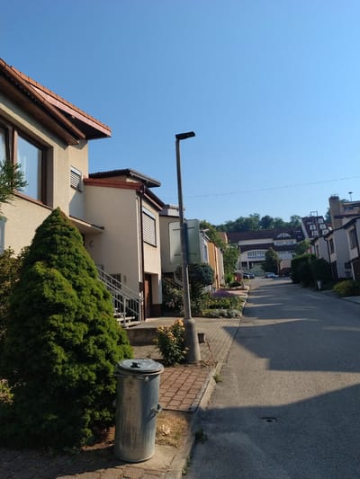 The image depicts a street scene in Hluboká nad Vltavou, Czechia. The perspective is from ground level, looking down a residential street lined with modern, multi-story houses. A large, conical evergreen tree is prominent in the foreground to the left, next to a silver, cylindrical trash bin. The street is paved and appears to be empty of people, though a few cars are visible parked further down the road and one driving away. The architecture suggests a suburban setting with a clear blue sky overhead. The houses have beige or white facades, some with brown tiled roofs and dark-framed windows, others with lighter colored elements. A street lamp stands on the right side of the street. The overall impression is a quiet, sunny day in a well-maintained neighborhood.