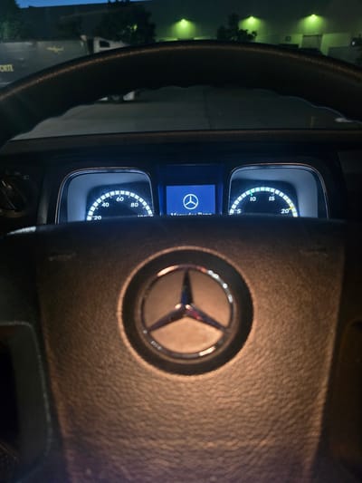 The image shows the interior of a Mercedes-Benz truck at night. The perspective is from the driver's seat, looking forward. The steering wheel, with the Mercedes-Benz logo prominently displayed, is in the foreground. Behind the steering wheel are two digital instrument clusters. The left cluster displays a speedometer with markings from 0 to 100, indicating a current speed of approximately 20. The right cluster shows a gauge with markings from 0 to 20, with a needle pointing between 15 and 20, possibly representing engine RPM or fuel level. A blue screen on the left displays the Mercedes-Benz logo. Outside the vehicle, dimly lit by green lights, are buildings and trees, suggesting an industrial or commercial area. The scene is captured in low light conditions, typical of nighttime. The location is identified as Zeppelinheim, Germany.