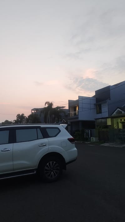 A white SUV is parked on an asphalt road in front of modern houses with gray and white facades, and a yellow house with a green fence. Palm trees are visible behind the SUV and between the houses. The sky is light gray with hints of pink and orange, suggesting it is either sunrise or sunset. The lighting is soft, and there are some faint clouds. The overall scene appears to be a residential area in Pusakarakyat, Indonesia, possibly during a quiet moment in the early morning or late evening. There is no visible text in the image.