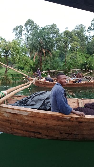 A man sits in a wooden boat on a body of water, possibly a lake or river. Other people are in the boat with him. The boat appears to be handmade, constructed from planks of wood. Trees and other foliage line the bank, suggesting a rural or natural setting. The water reflects the green hues of the surroundings.