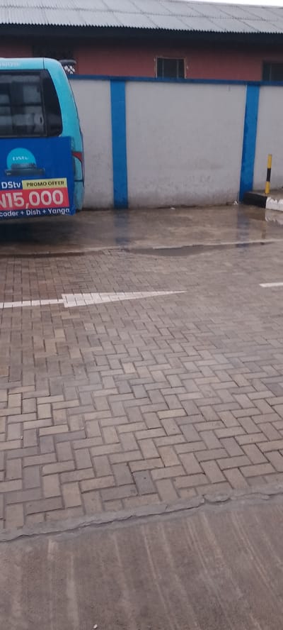 The image shows a blue and teal colored bus parked on a wet, paved ground. The bus has text on its side advertising a "DStv PROMO OFFER" for "N15,000" which includes "decoder + Dish + Yanga". The background consists of a light-colored wall with blue stripes, and a corrugated metal roofed building. There is a yellow and black bollard on the right side of the image, near the wall. The ground is made of interlocking pavers and appears wet, suggesting recent rain or damp conditions. The overall scene suggests an outdoor commercial or transportation hub in Jibowu, Nigeria, possibly during a cloudy or overcast day. There are no people visible in the image.