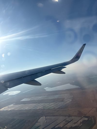 The image displays an aerial view of a landscape identified as Nezahualcóyotl, Mexico, seen from an aircraft in flight. A prominent feature is the dark gray aircraft wing, bearing the "volaris.com" branding on its upward-curved wingtip, extending from the middle-left towards the upper-right of the frame. The sky above is clear blue, with bright sunlight emanating from the upper-left, creating visible lens flare and circular light artifacts.
Below the wing, the terrestrial environment consists of a flat, semi-arid terrain characterized by numerous man-made modifications. Several irregularly shaped, shallow water bodies are present, reflecting the sky. Large, distinct rectangular and linear patterns are visible across the land, suggesting structured land use such as agricultural fields, water retention basins, or potentially salt evaporation ponds. The ground itself is predominantly brown and gray, with visible variations in texture and moisture content. In the far distance, towards the right side of the horizon, a hazy outline of urban development or distant structures can be discerned. No individuals or specific human activity are visible within the frame. The scene is illuminated by daylight, under clear atmospheric conditions with some visible haze affecting distant views.