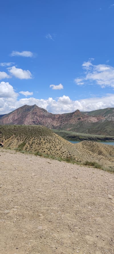 The image depicts a mountainous landscape under a blue sky dotted with cumulus clouds. A large, reddish-brown mountain range dominates the midground, with patches of green vegetation visible on its slopes. In front of the mountains, there's a series of rolling hills covered in sparse, dry vegetation. A body of water, likely a lake or reservoir, is nestled between the hills. The foreground is composed of rocky, dirt terrain. The scene appears to be in a rural location, potentially near Lanjazat, Armenia. No people are visible in the frame.