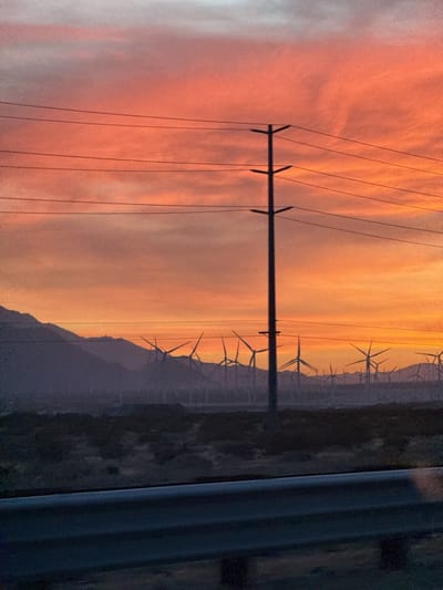 The image depicts a landscape near Desert Hot Springs, United States, at sunset. The sky is filled with orange and pink hues. An electrical transmission tower stands prominently in the foreground, with multiple power lines stretched horizontally across the frame. Behind the tower, a large field of wind turbines are visible against the backdrop of mountains. A dark, low-lying scrubland lies in front of the turbines. A vehicle's guardrail is visible in the lower foreground of the image, suggesting the picture was taken from a moving vehicle.